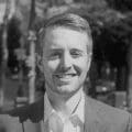 A grayscale photo of a man with short hair, wearing a suit and a white dress shirt. He is smiling and positioned outdoors with trees and other blurred background elements. The overall atmosphere suggests a pleasant day.