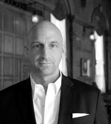 A bald man in a suit and white shirt stands in a dimly lit, elegant room with wooden paneling and arched windows. He has a slight smile and a white pocket square visible in his blazer. The background features ornate decor and framed pictures.