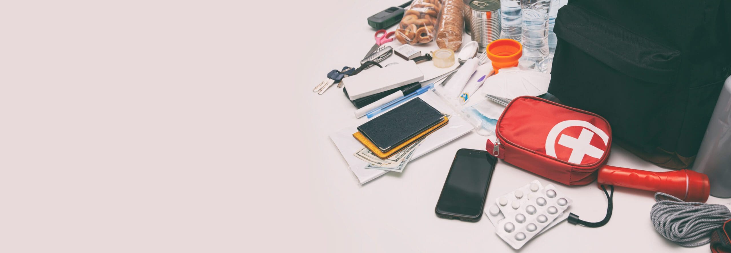 A variety of emergency kit items are spread out on a white surface. Visible items include a red first aid kit, flashlight, medicine, smartphone, notebook, keys, canned goods, snacks, water bottles, and other essentials. A black backpack is seen on the edge.